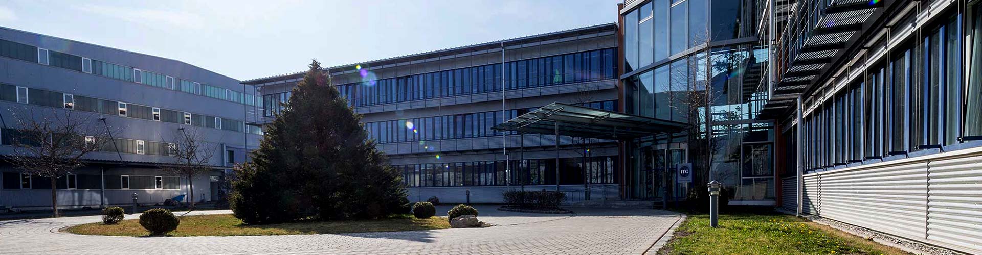 Exterior view of a modern office and administrative building with glass facade, entrance area, and paved forecourt